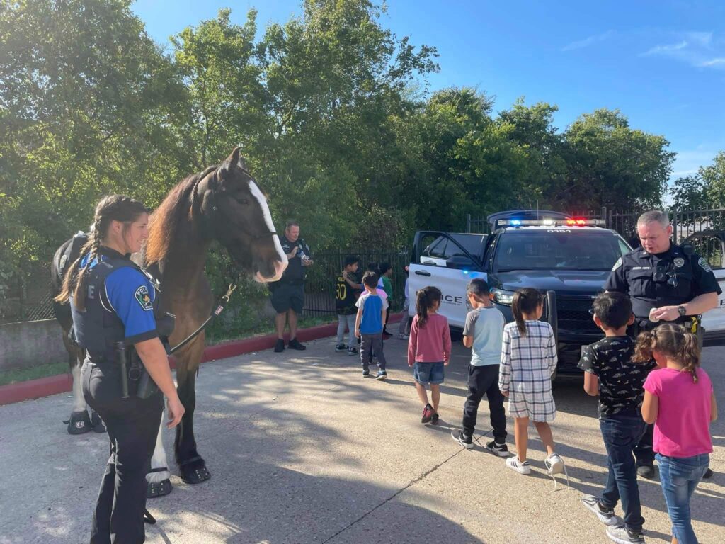 Police officers interacting with children near a patrol car and a mounted officer standing next to a horse.