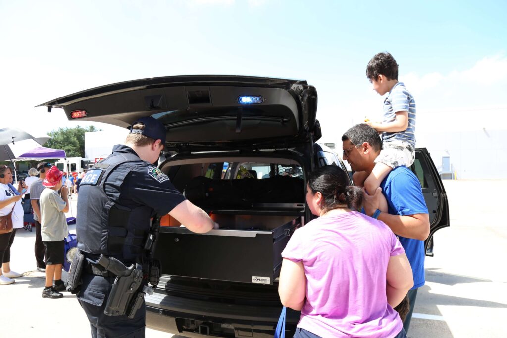 A police officer showing a patrol car's trunk to a family with a child sitting on an adult's shoulders.