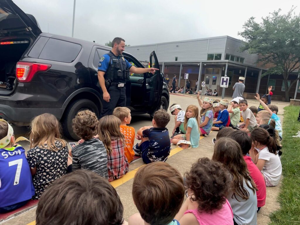 A police officer speaking to seated children in front of a police SUV on Career Day.