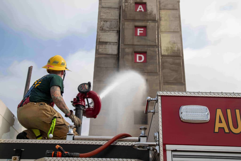 A firefighter in gear operating a hose from atop fire truck, spraying water at a tall training tower in the background.