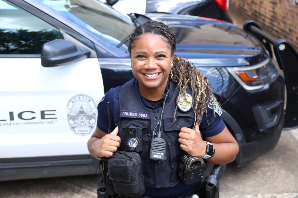 A smiling police officer standing in front of a police car holding her vest straps.