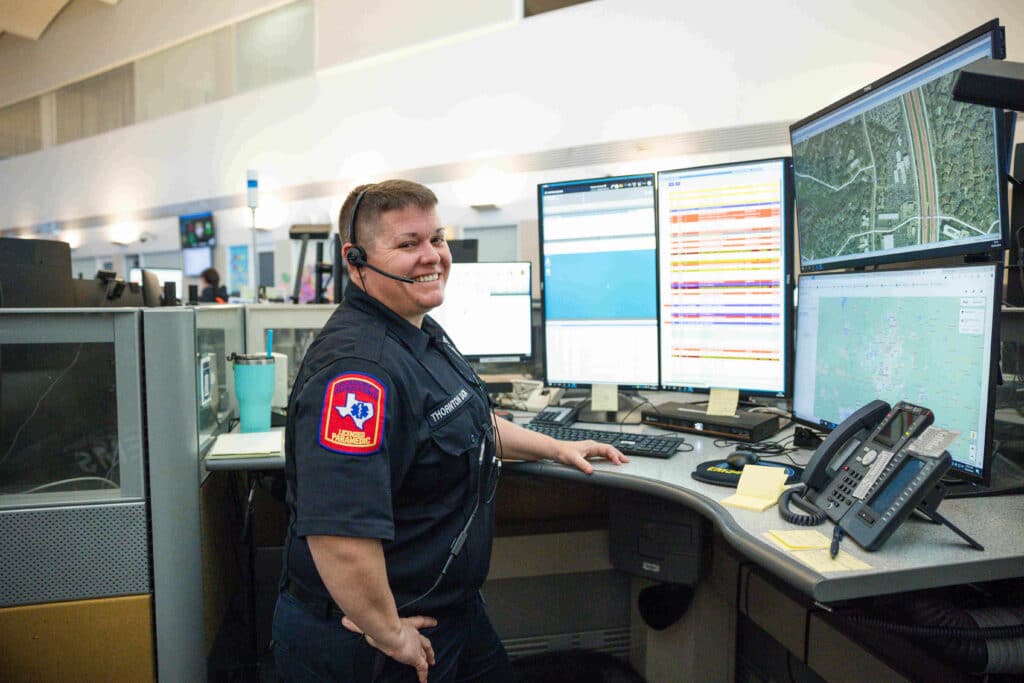 A smiling paramedic wearing a headset and uniform in a dispatch center, surrounded by multiple monitors.